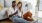 a man and woman with take out, sit against a couch on their living room floor and watch at their cell phone